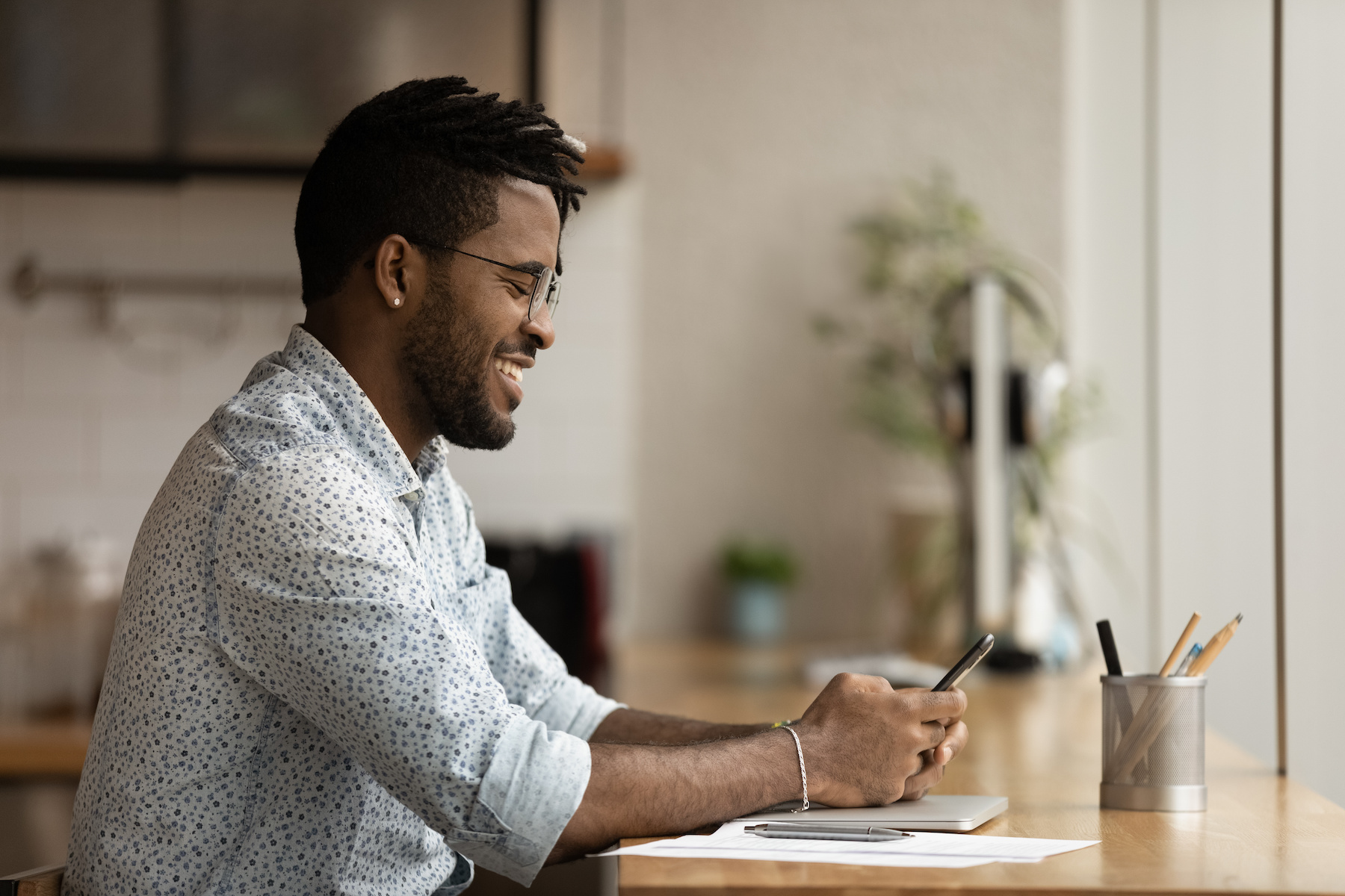 Young african mixed race man using cellphone applications, distracted from computer work in modern office, communicating online, checking email, relaxing watching funny video or shopping.