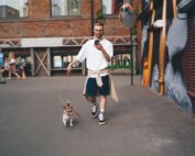 Young male in casual clothes text messaging on cellphone while walking with small Jack Russell Terrier dog along street in city.
