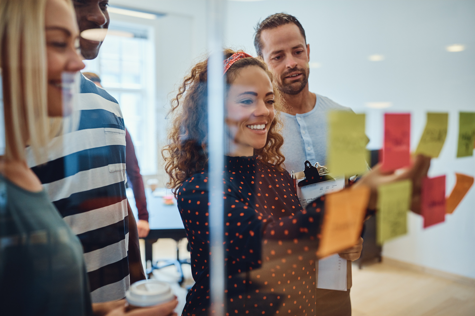 Smiling group of designers reading sticky notes on a glass wall during a brainstorming session.