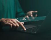 A person wearing a green long sleeve shirt, holding a cellphone and typing on a laptop with a graphic of a semi transparent internet search bar overlayed