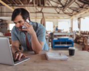 Man leaning on table while talking on the phone and using laptop in workshop
