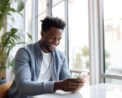 Man smiling at phone while sitting down on table next to window.