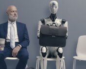 Man wearing suit and sittting on chair next to robot holdiing a brief case.