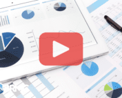 A close-up of a tablet displaying various financial charts, including pie charts and line graphs, alongside printed spreadsheets on a desk. A pen is placed on the papers, and a large red play button icon is overlaid, indicating video content. The BRMC logo, featuring a mountain design, is visible in the bottom-left corner.