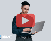 A man in a dark blue shirt uses a laptop while looking focused, with a play button overlay centered on the image. The BRMC logo is visible in the bottom left corner.