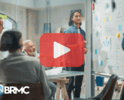 A group of people is gathered in a modern meeting room with glass walls. In the foreground, a man with shoulder-length dark hair stands by a whiteboard covered with colorful sticky notes and hand-drawn diagrams. Seated nearby, an older man with white hair and a beard looks on attentively, joined by other colleagues. The atmosphere is collaborative and focused, reflecting a professional brainstorming session. A semi-transparent red "play" button is overlaid in the center, indicating a video or presentation context. The BRMC logo, with a mountain peak icon, is visible in the bottom left corner.