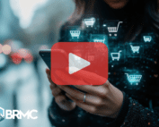 A person using a smartphone for online shopping, with futuristic shopping cart icons overlaying the image, representing digital commerce. The BRMC logo and a play button suggest video content.