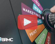 A close-up image of a hand turning a dial labeled with various advertising and marketing categories such as “Marketing,” “Web,” “Print,” and “Design.” A large translucent play button is overlaid on the image, suggesting it's a video thumbnail. A logo labeled "BRMC" is in the bottom left corner.