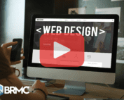 A person is sitting at a desk in front of a large desktop monitor displaying a website labeled “WEB DESIGN.” A smartphone is in one hand, and a large play button overlays the screen, suggesting video content. The BRMC logo is visible in the corner.