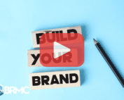 A set of wooden blocks arranged to spell out the words "BUILD YOUR BRAND." A pencil sits next to the blocks on a light blue background, symbolizing the concept of constructing or developing a brand.